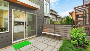 Une maison moderne avec une entrée dotée d’une porte en verre, qui donne sur une cuisine, un banc en bois et un petit espace jardin.