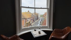 A room with a large window offering a view of a street. There is a small table with two chairs and two glasses of wine on it.