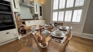 A modern kitchen with a glass dining table set for breakfast, wooden flooring, and a large window with blinds.