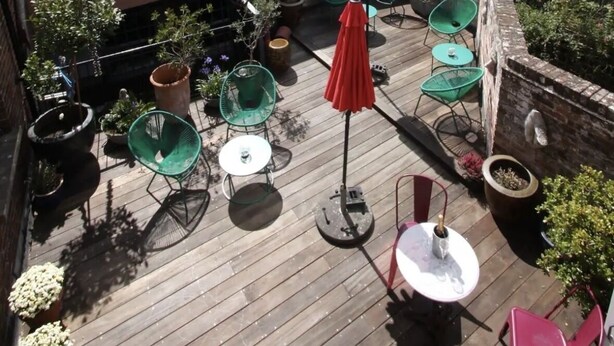 A wooden deck with outdoor seating, including tables and chairs, surrounded by potted plants and a red umbrella.