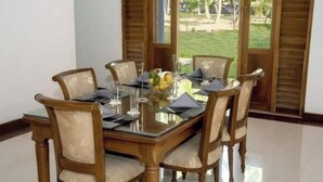 A dining area with a glass-top table, wooden chairs, and a view of a garden through large windows. - Coco Villa Peach Tree (Seeduwa - Katunayake)