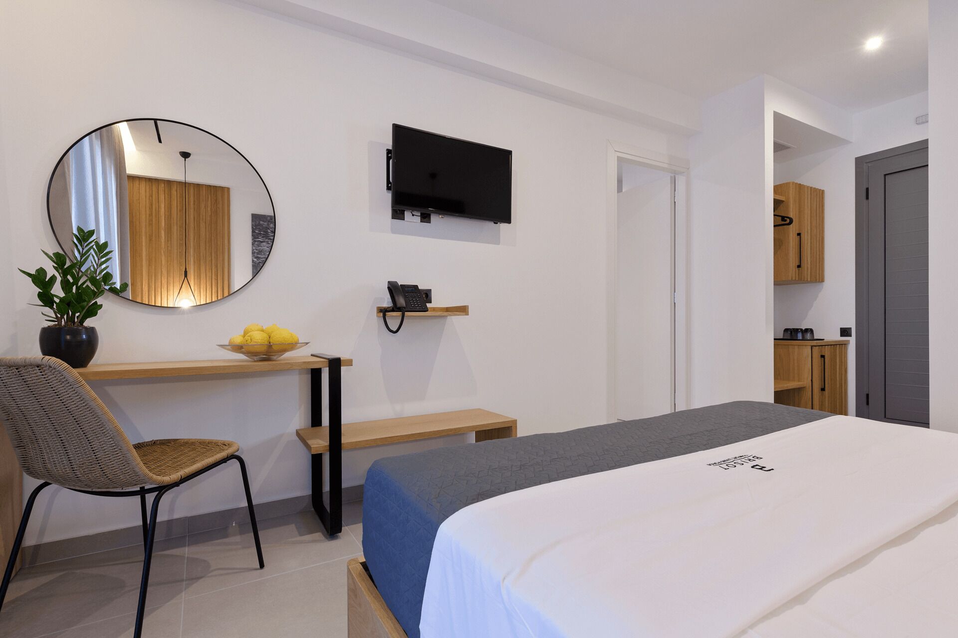 A modern hotel room with a bed, a desk with a chair, a TV, and a small kitchen area.