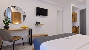 A modern hotel room with a bed, a desk with a chair, a TV, and a small kitchen area.
