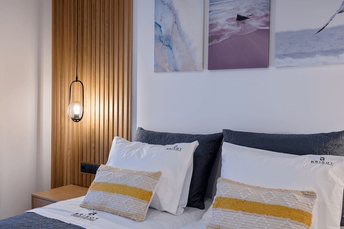 A neatly made bed with two pillows, a bedside table, a wall-mounted lamp, and two framed artworks.