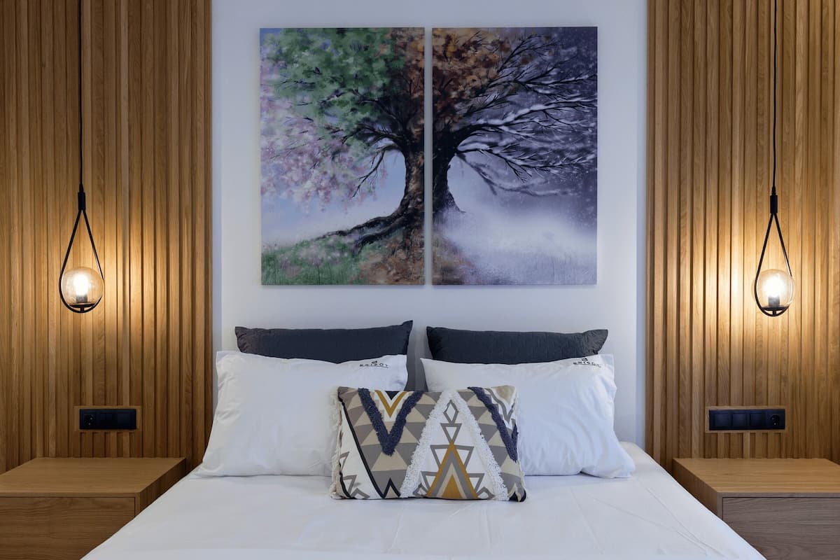 A bedroom with a bed, two wall-mounted lamps, and two paintings of trees.