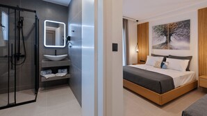 A modern bathroom with a glass shower enclosure, a white sink, and a mirror with integrated lighting. The bedroom features a large bed with a gray and white bedding set, a wooden headboard, and a painting of a tree on the wall. - BRISOT Hotel Galatas Poros (Poros)