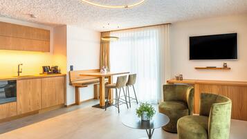 A modern living room with a round coffee table, two green armchairs, a dining area with a wooden table and chairs, and a kitchen with wooden cabinets and a built-in oven.