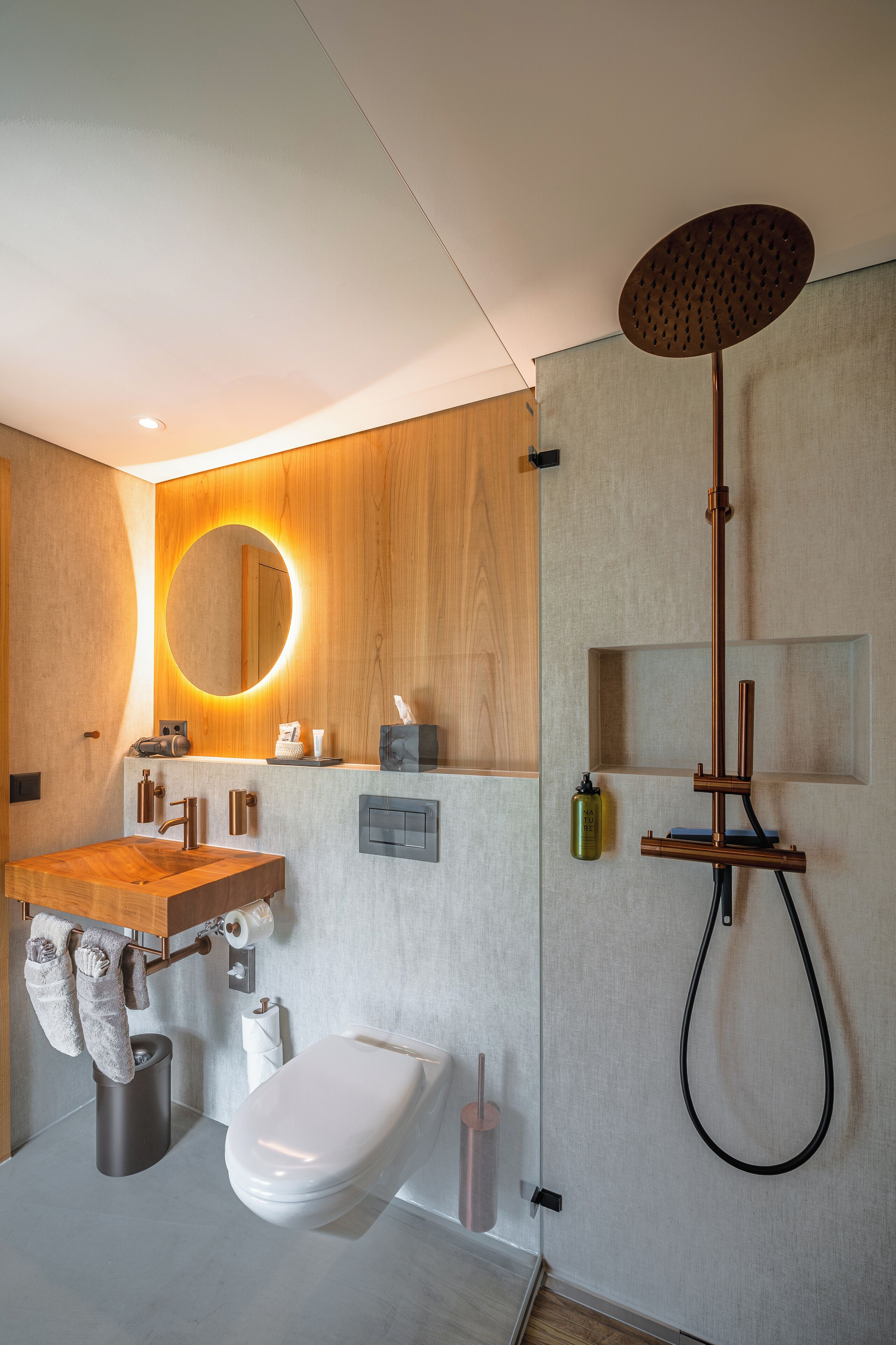 A modern bathroom with a wooden vanity, a round mirror, a toilet, and a shower with a unique headrest.