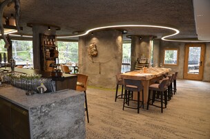 Um restaurante com interior rústico, apresentando uma parede de pedra, mobiliário de madeira e um espaço de bar com copos e garrafas.