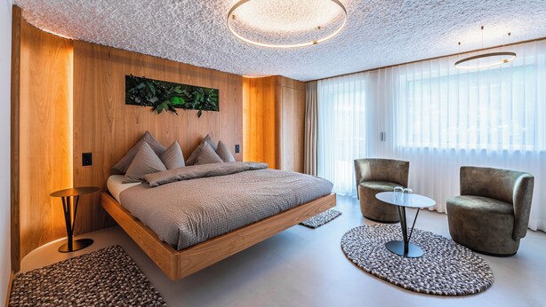A modern bedroom with a floating bed, a round table, two armchairs, and a large window with sheer curtains. - Boutique Hotel La Gorge (Saas-Fee)