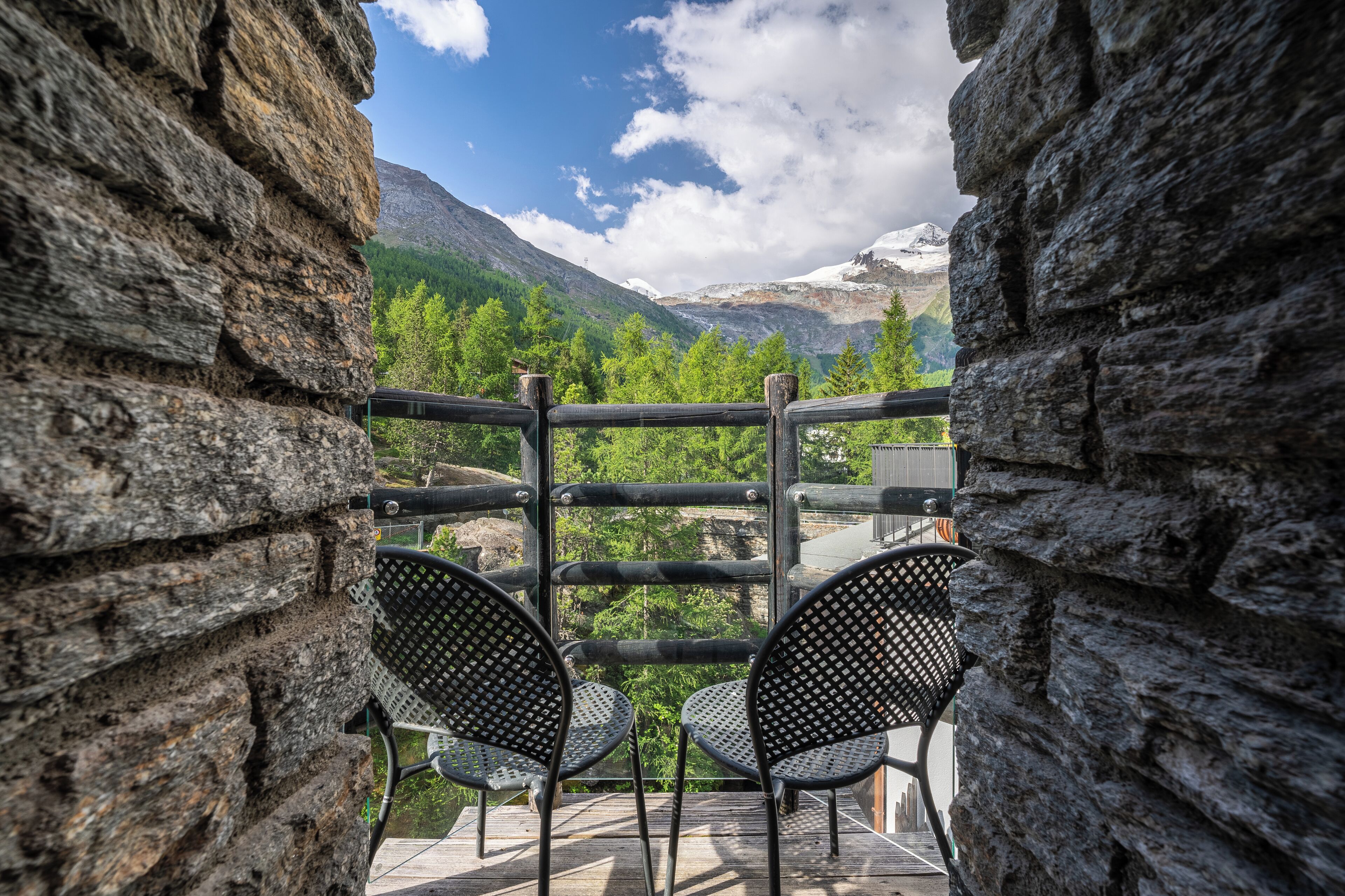 Ein Balkon mit zwei Stühlen, einer Steinmauer und Blick auf Berge und Bäume.