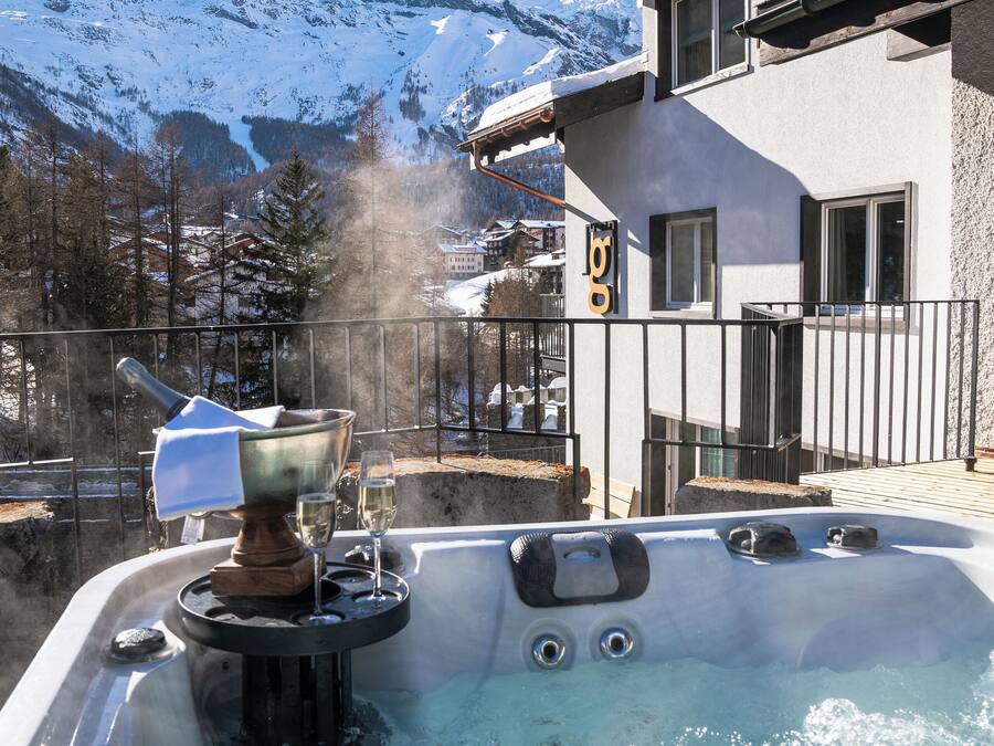 Un jacuzzi con vistas a montañas nevadas, un edificio de estilo chalet y vapor ascendiendo.