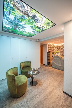 Un interior moderno con una gran pantalla digital mostrando un paisaje natural, dos sillones verdes, una mesita redonda y una pared de piedra con un estante.
