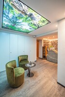 A modern interior with a large digital screen displaying a natural scene, two green armchairs, a small round table, and a stone wall with a shelf.