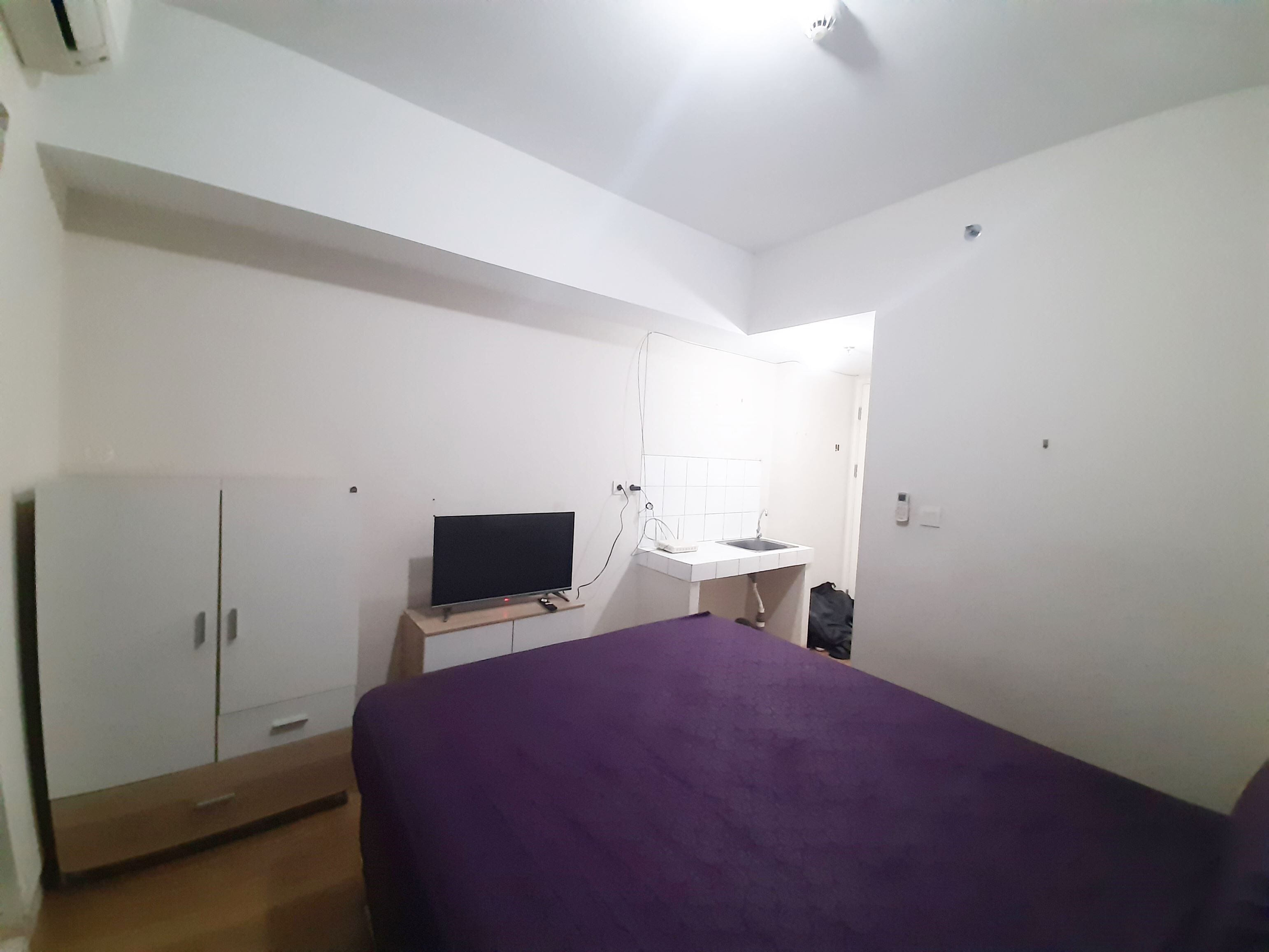 A bedroom with a bed, a TV on a stand, a wardrobe, and a bathroom visible through an open door.