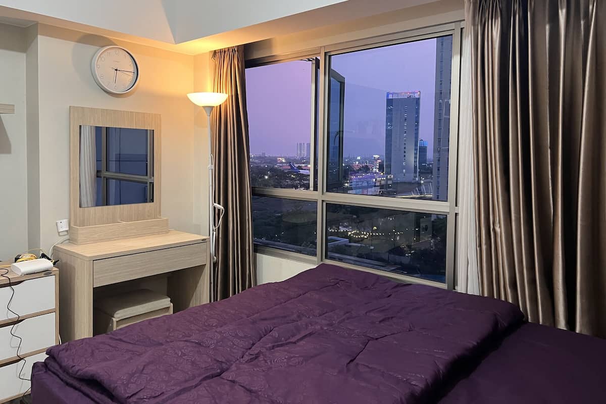 A hotel room with a bed, a desk, a chair, a lamp, a clock, and a window with curtains.