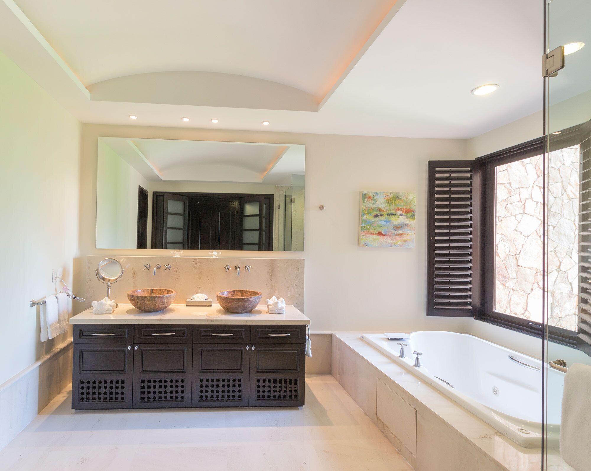 A modern bathroom with a large mirror, two sinks, a bathtub, and a window with blinds.