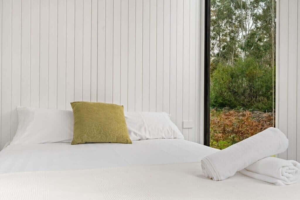 A neatly made bed with white linens and a single green pillow, a white towel on the floor, and a window with a view of trees and foliage.