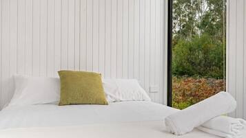 A neatly made bed with white linens and a single green pillow, a white towel on the floor, and a window with a view of trees and foliage.