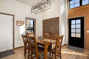 Zona pranzo con tavolo e sedie in legno, una lampada a sospensione e ampie finestre.