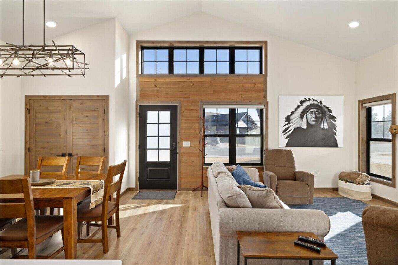 A spacious living area with a high ceiling, wooden paneling, a dining table with chairs, a sofa, and a large window with a view.