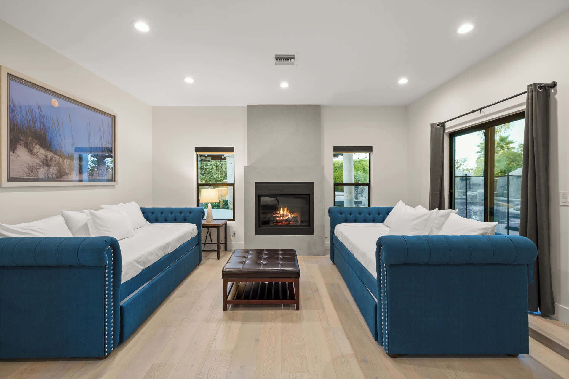 A modern living room with two blue sofas, a central fireplace, and large windows offering views of greenery.