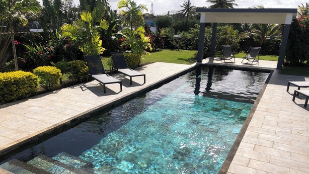 A rectangular swimming pool with clear water, surrounded by a tiled deck. There are lounge chairs and a covered seating area with a pergola.