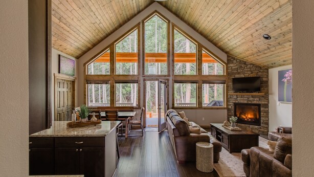 A spacious loft with a high ceiling, large windows offering a view of the forest, a fireplace, and a cozy seating area.