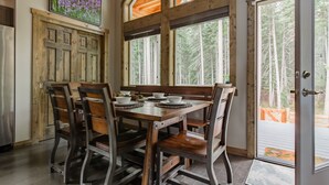 A dining area with a wooden table and chairs, large windows with a forest view, and a door leading outside.