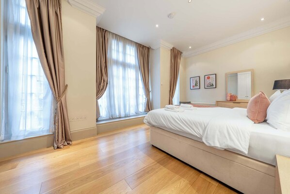 Een ruime slaapkamer met een groot bed, houten vloeren en grote ramen voorzien van gordijnen.