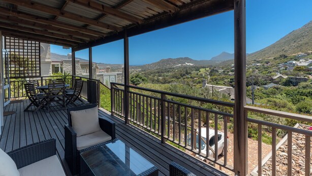Una terraza de madera con mobiliario exterior, con vistas a un paisaje montañoso con casas.