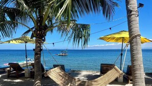Escena de playa con una hamaca, sillas de descanso y un barco al fondo.