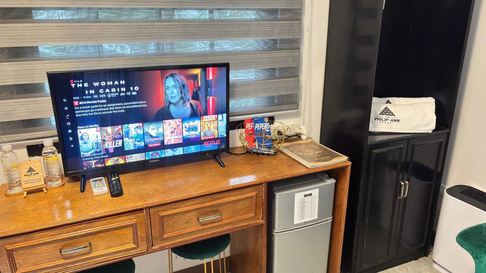 A hotel room with a wooden desk, a flat-screen TV displaying a movie, a minibar, and a minibar fridge.