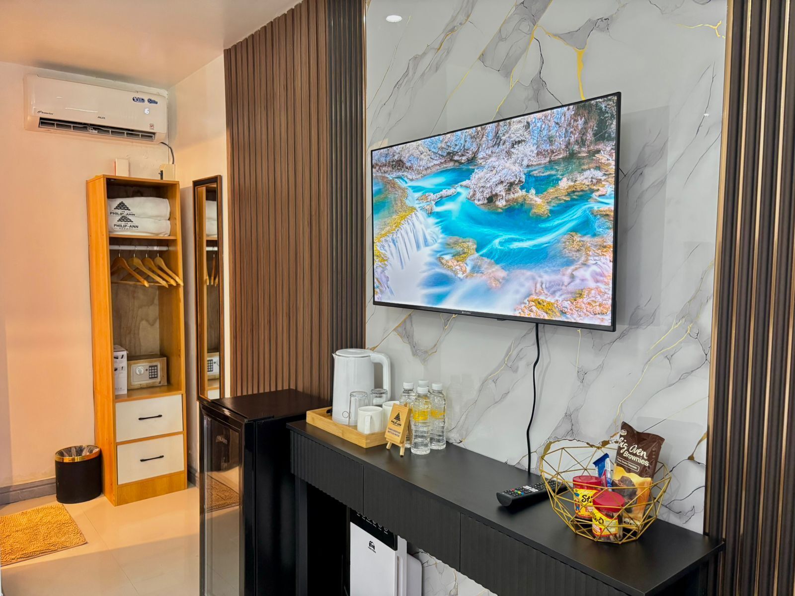 Habitación de hotel moderna con pared de mármol, televisor montado, armario de madera, estación de café y minibar.