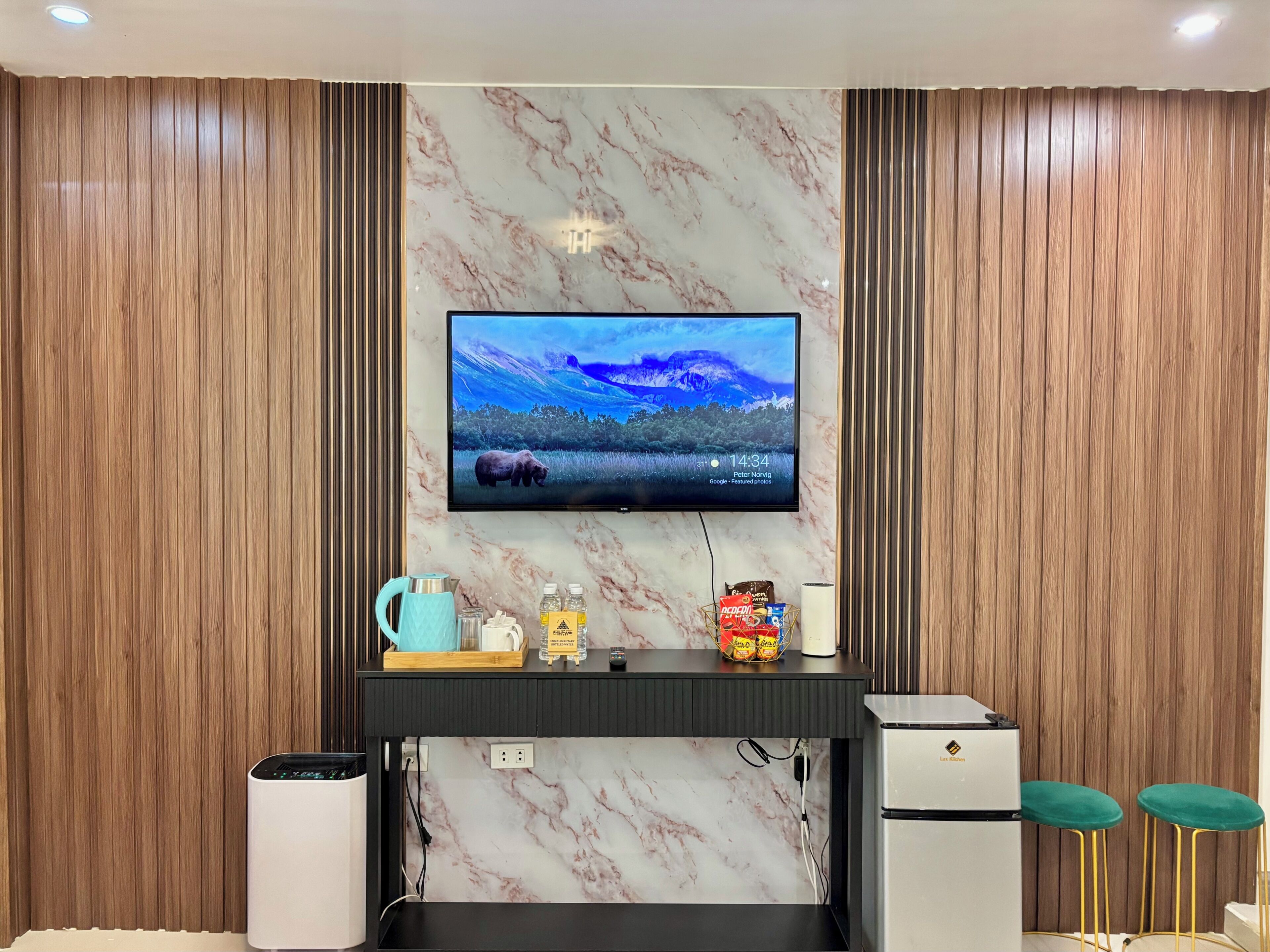 A modern living room with a flat-screen TV mounted on a marble wall, a coffee maker, snacks, and a small refrigerator.