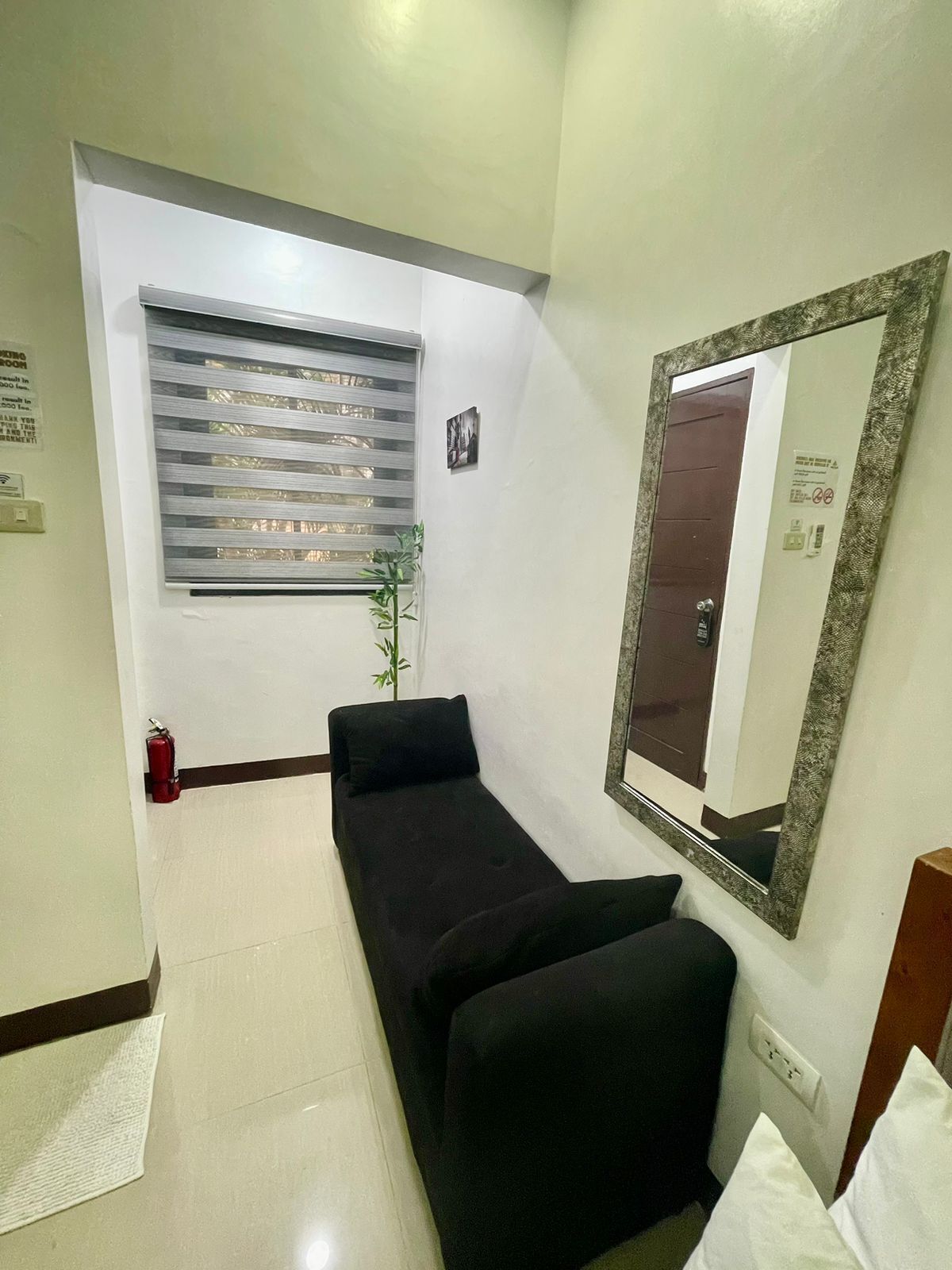 Una habitación pequeña y moderna con un sofá negro, una ventana con persianas, un espejo y un extintor.