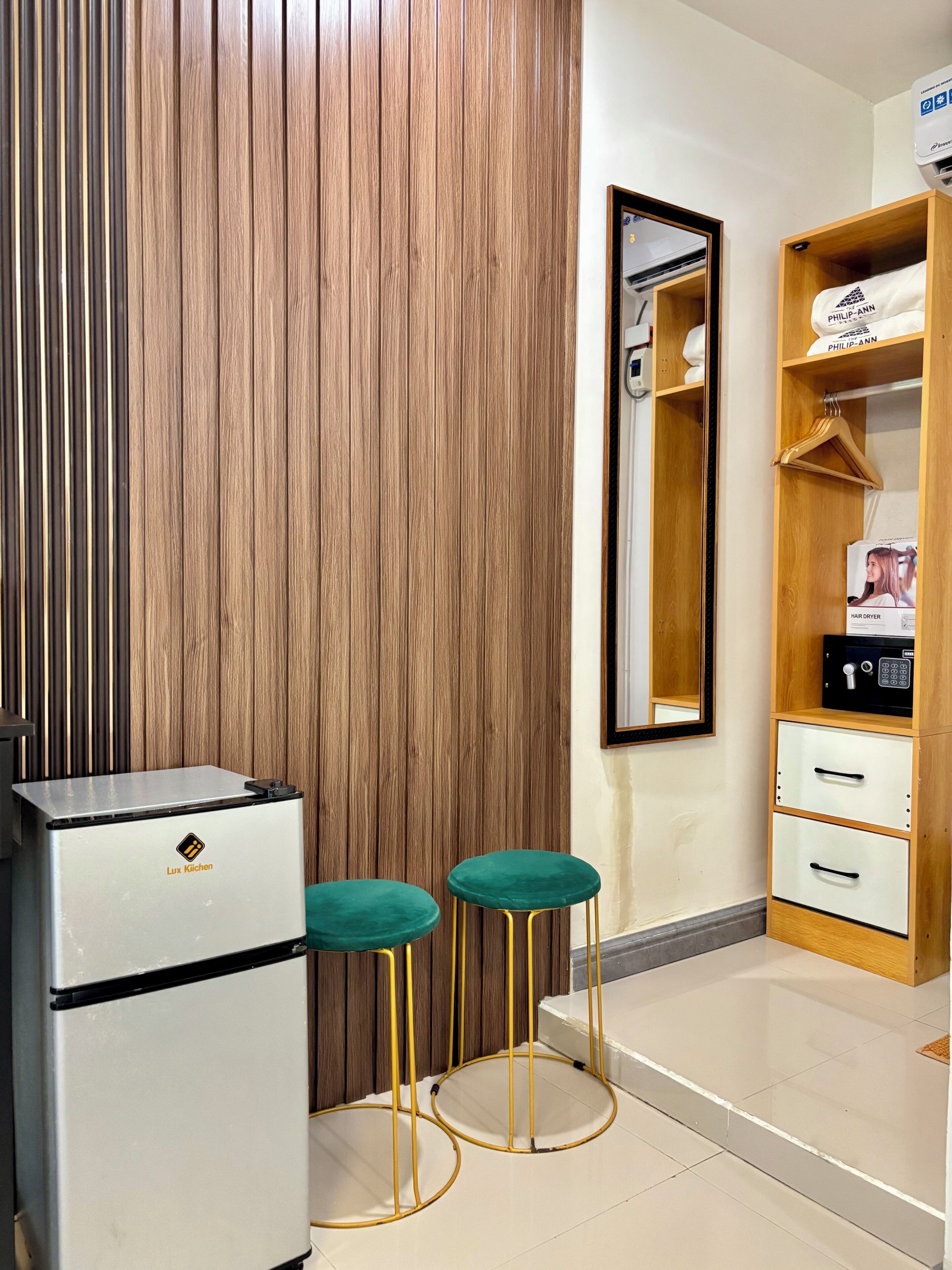 Quarto de hotel compacto com minibar, espelho, guarda-roupa e dois taburetes verdes.
