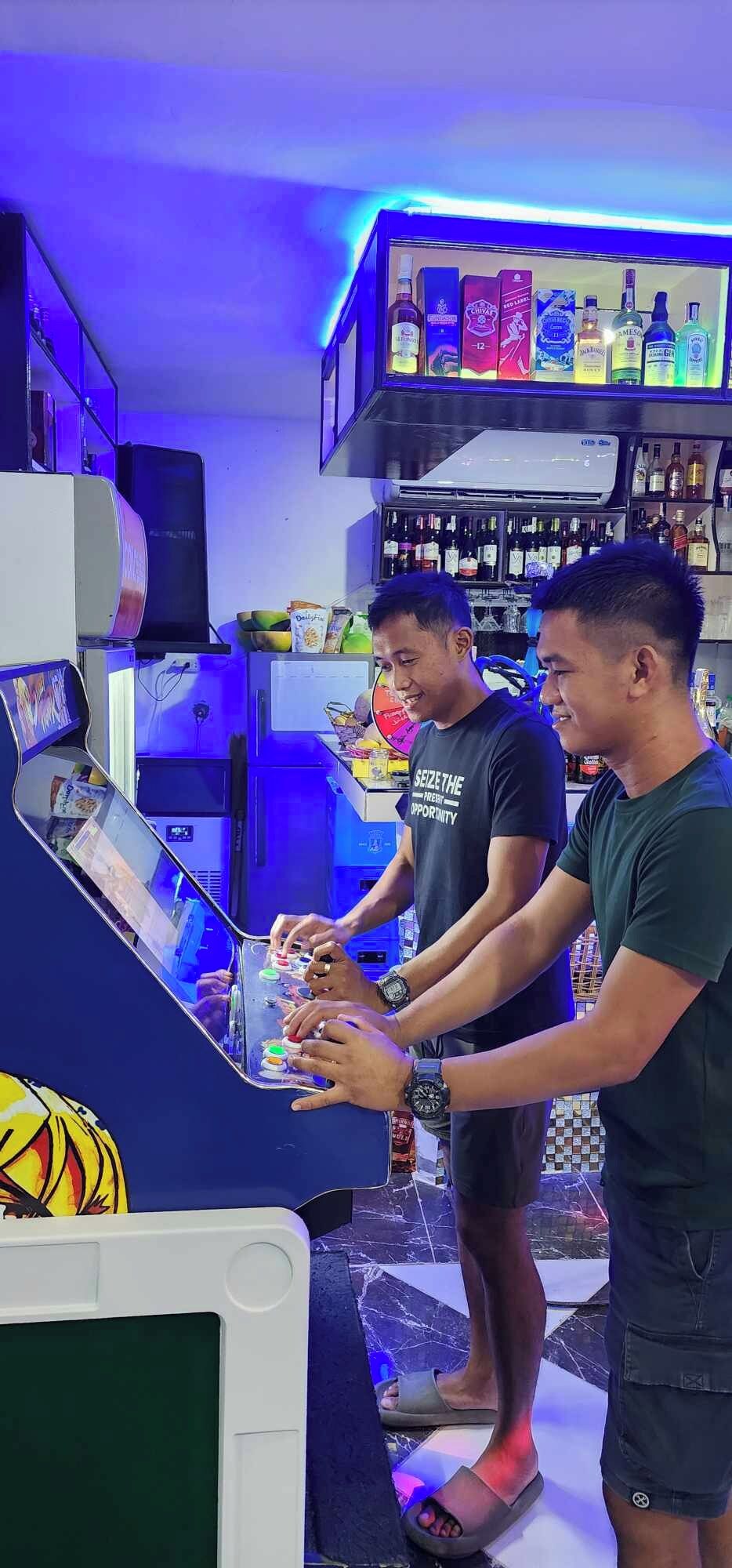 Un área de bar con mostrador, estantes llenos de botellas y dos personas de pie.