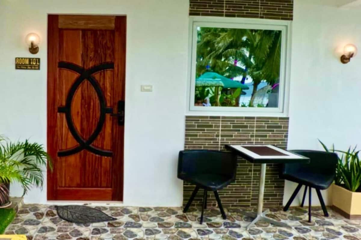 A room with a wooden door, a window with a view of palm trees, two chairs, and a small table.