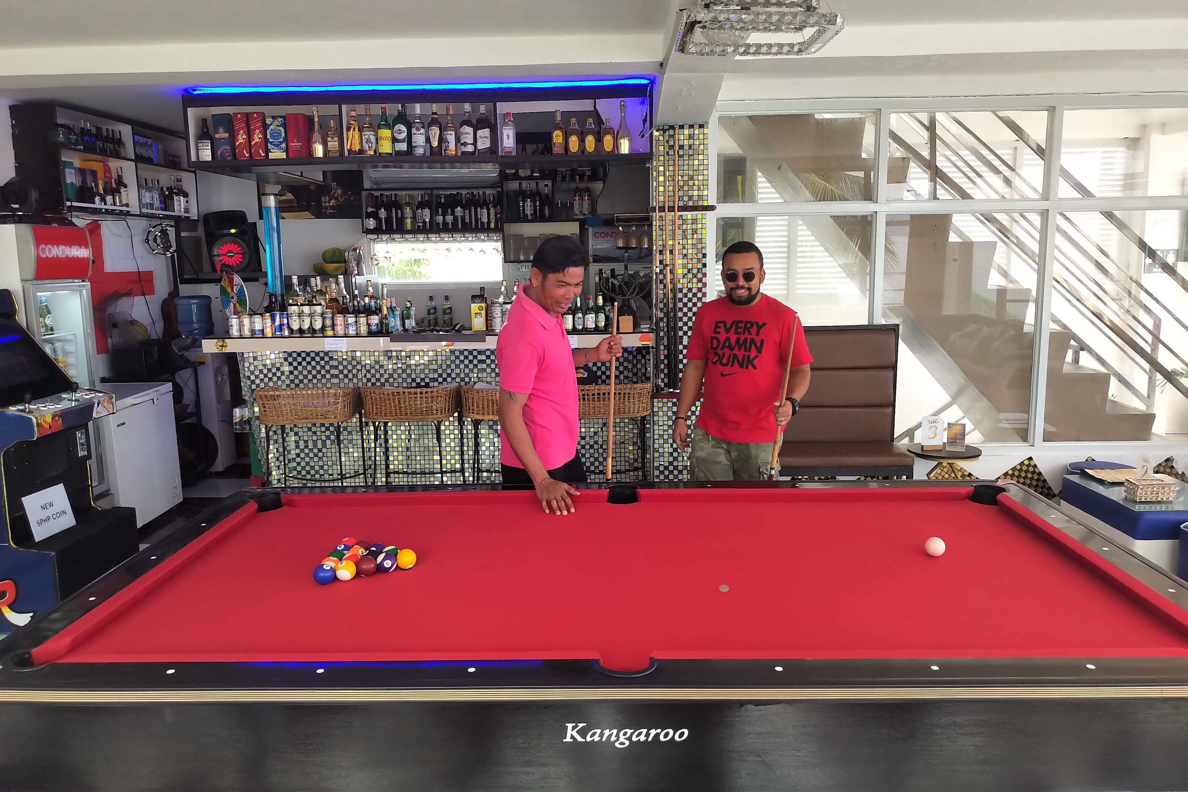 Une table de billard avec des billes, un bar avec différentes bouteilles, et deux personnes debout près de la table de billard.