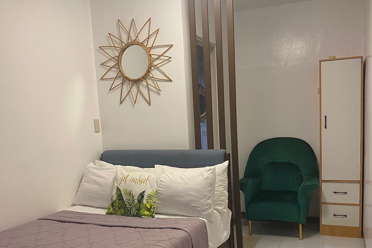 A bedroom with a bed, a green chair, a wardrobe, and a wall-mounted sun decoration.