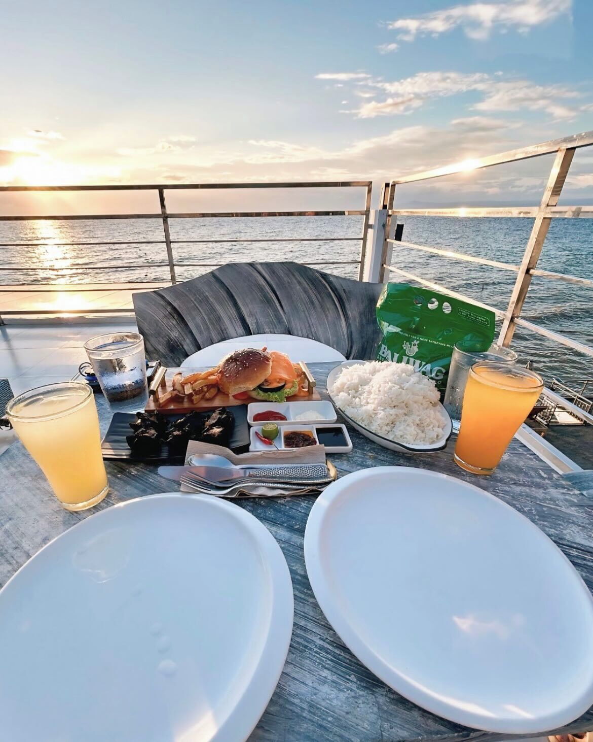 Une table à manger avec des assiettes, un hamburger, du riz et des boissons, le tout installé sur le pont d’un bateau, avec vue sur l’océan.
