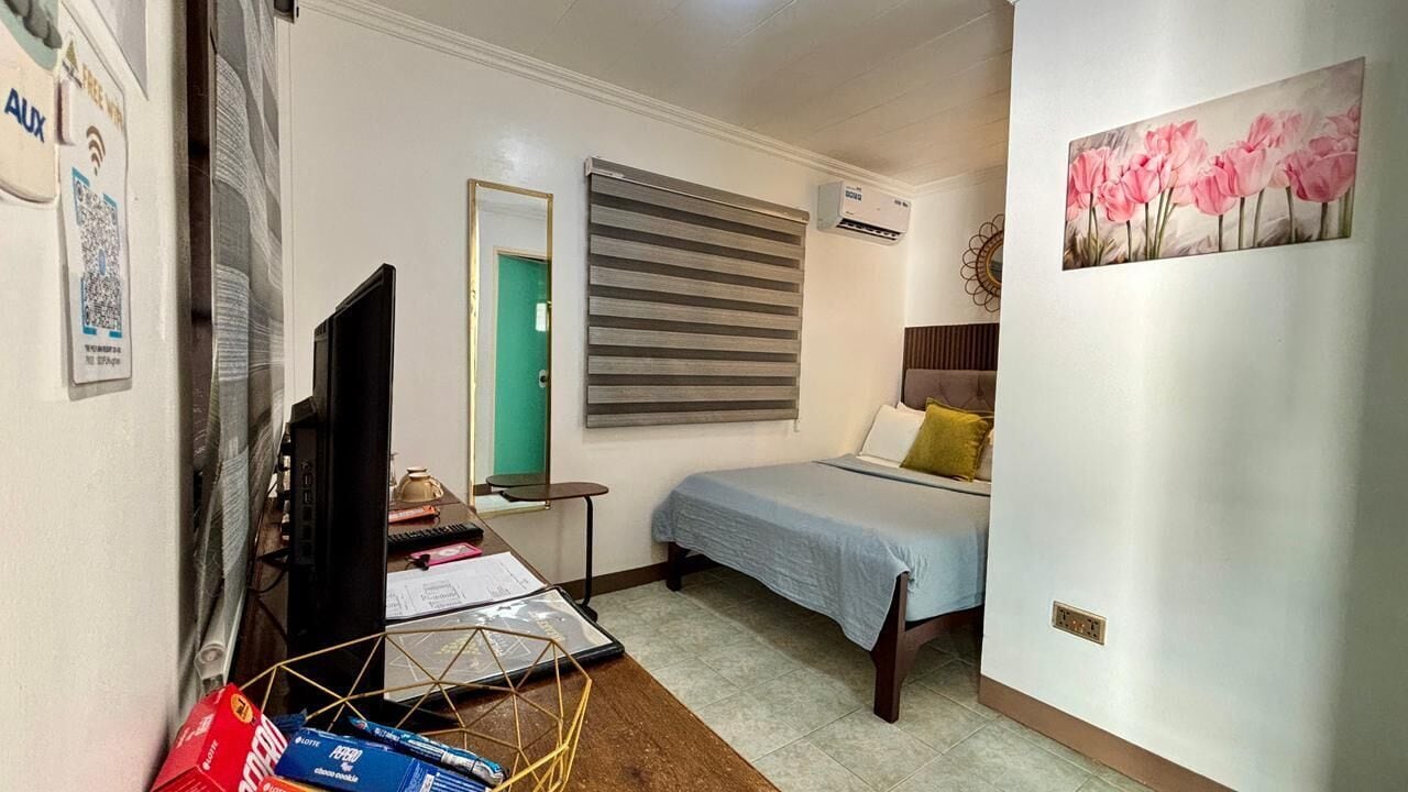 A compact hotel room with a bed, a desk with a computer, a mirror, and a painting on the wall.