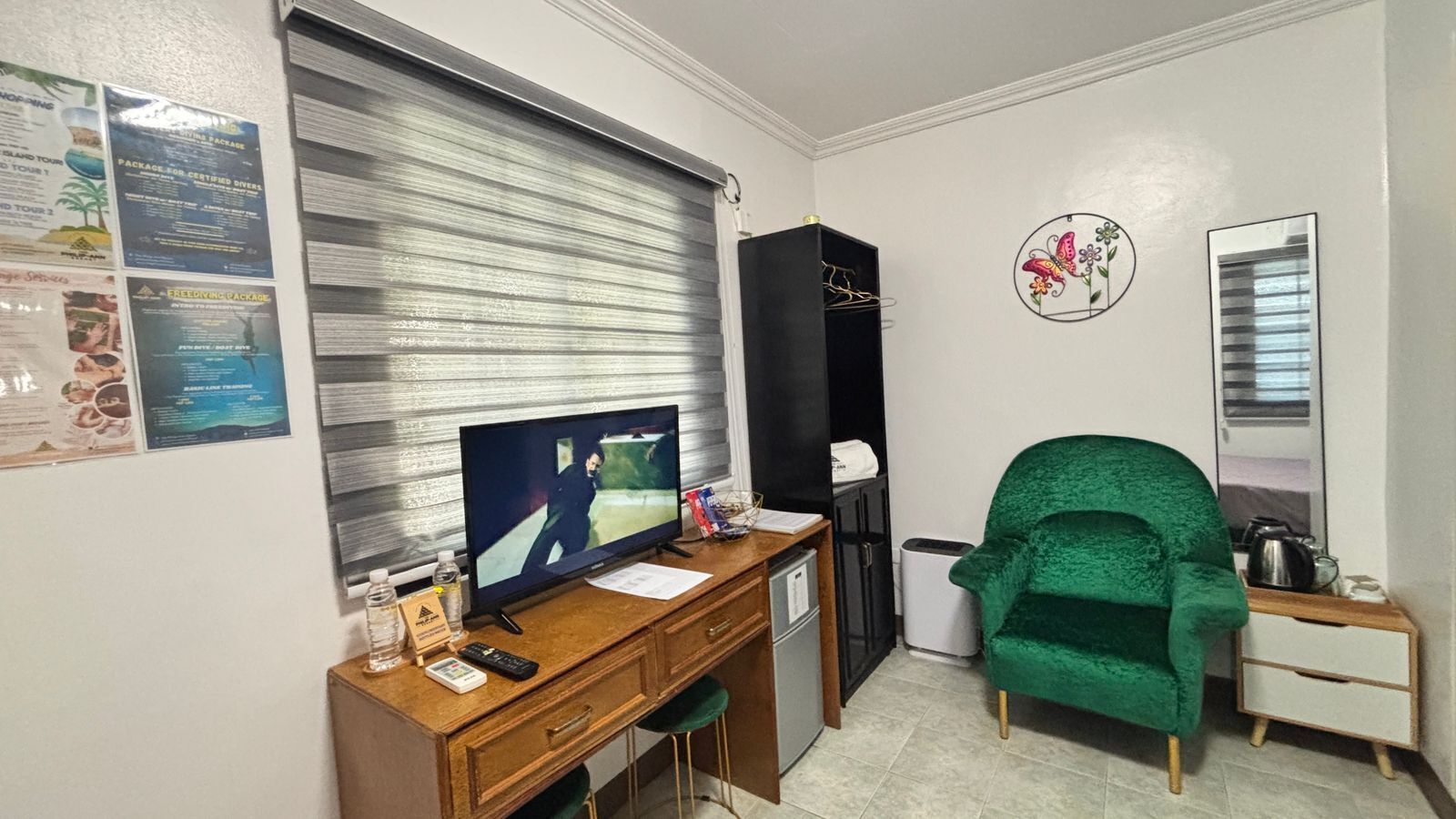 A room with a wooden desk, a television, a green chair, a black cabinet, and a wall with posters.