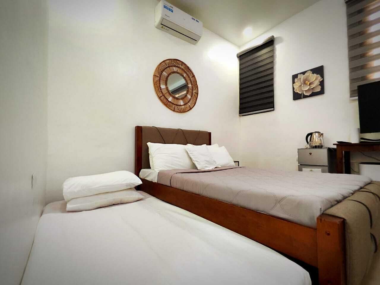 Une chambre avec deux lits, un miroir rond, un climatiseur fixé au mur, une fenêtre avec des stores et une petite table avec une lampe.