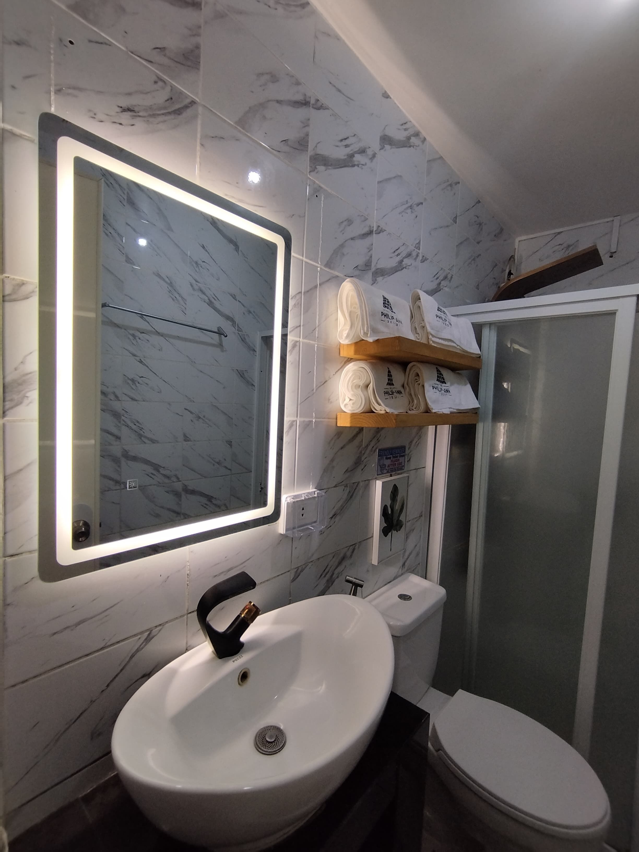 Een badkamer met een marmeren wand, een witte wastafel, een toilet, een douchecabine van glas en een plank met handdoeken.