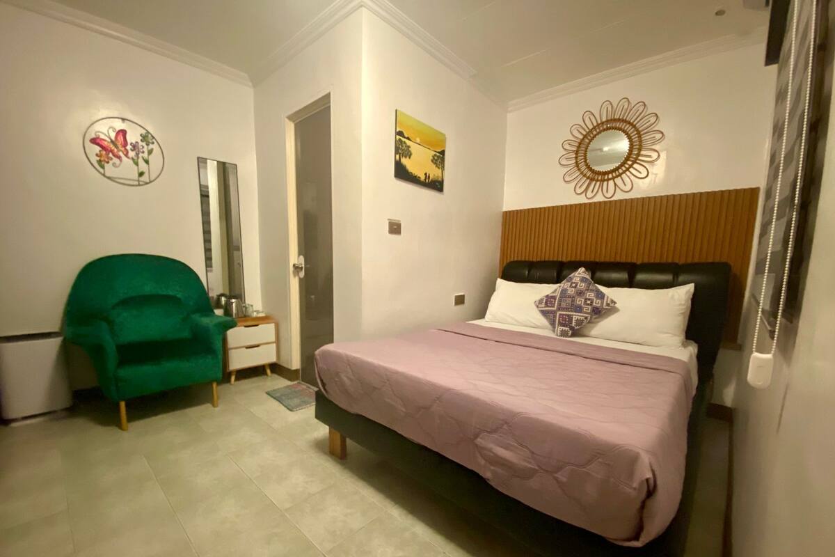 A bedroom with a bed, a green chair, a nightstand, a mirror, and a wall with a floral decoration.