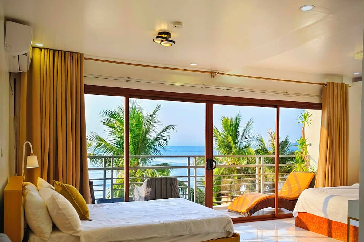 A modern hotel room with a large bed, a sofa, and a balcony with a view of the ocean and palm trees.