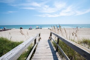Una pasarela de madera que lleva a una playa arenosa con sombrillas y gente.