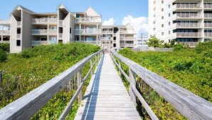 Une promenade en bois mène à un immeuble d’appartements situé en bord de mer, doté de nombreux balcons et offrant une vue dégagée sur l’océan.